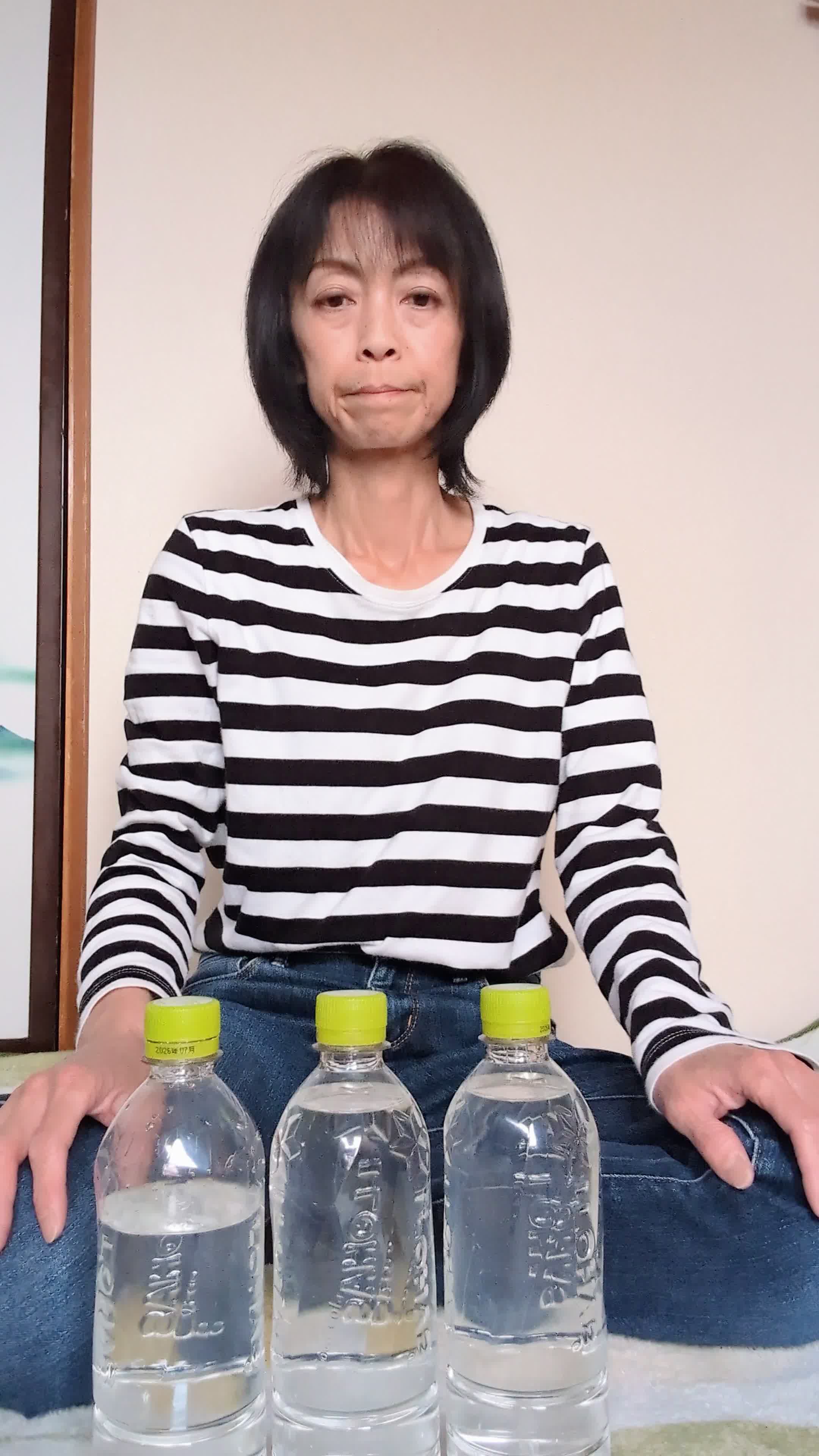 床に座り込み、スーパーの水のボトルを並べて「これを買い続けるのは大変」とため息をつく表情