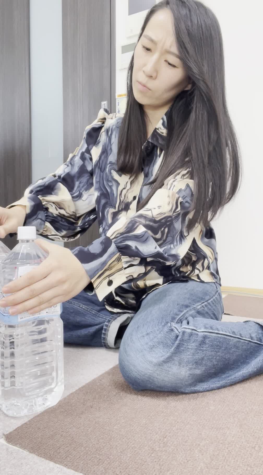床に座り込み、スーパーの水のボトルを並べて「これを買い続けるのは大変」とため息をつく表情