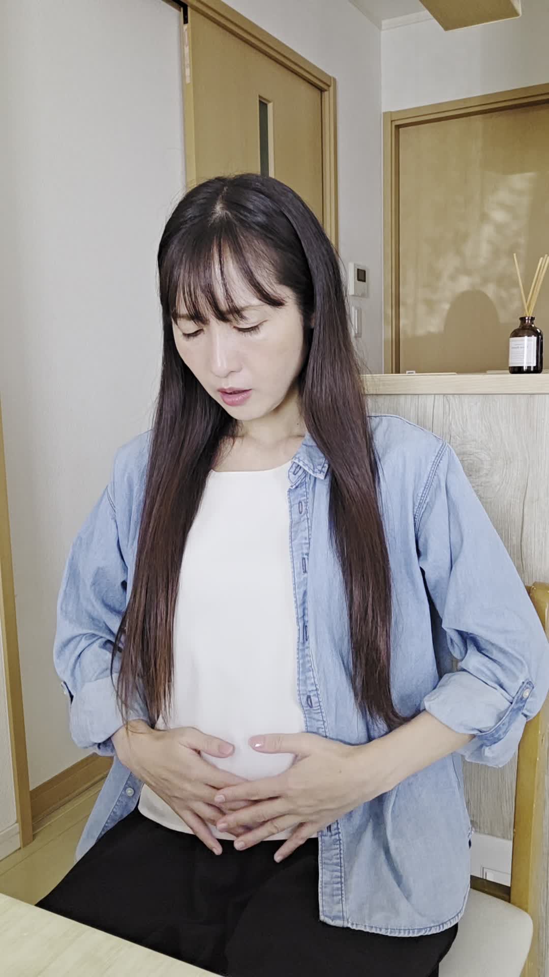 椅子に浅く腰掛け、「下腹部を両手で押さえて痛みを和らげようとする」動作