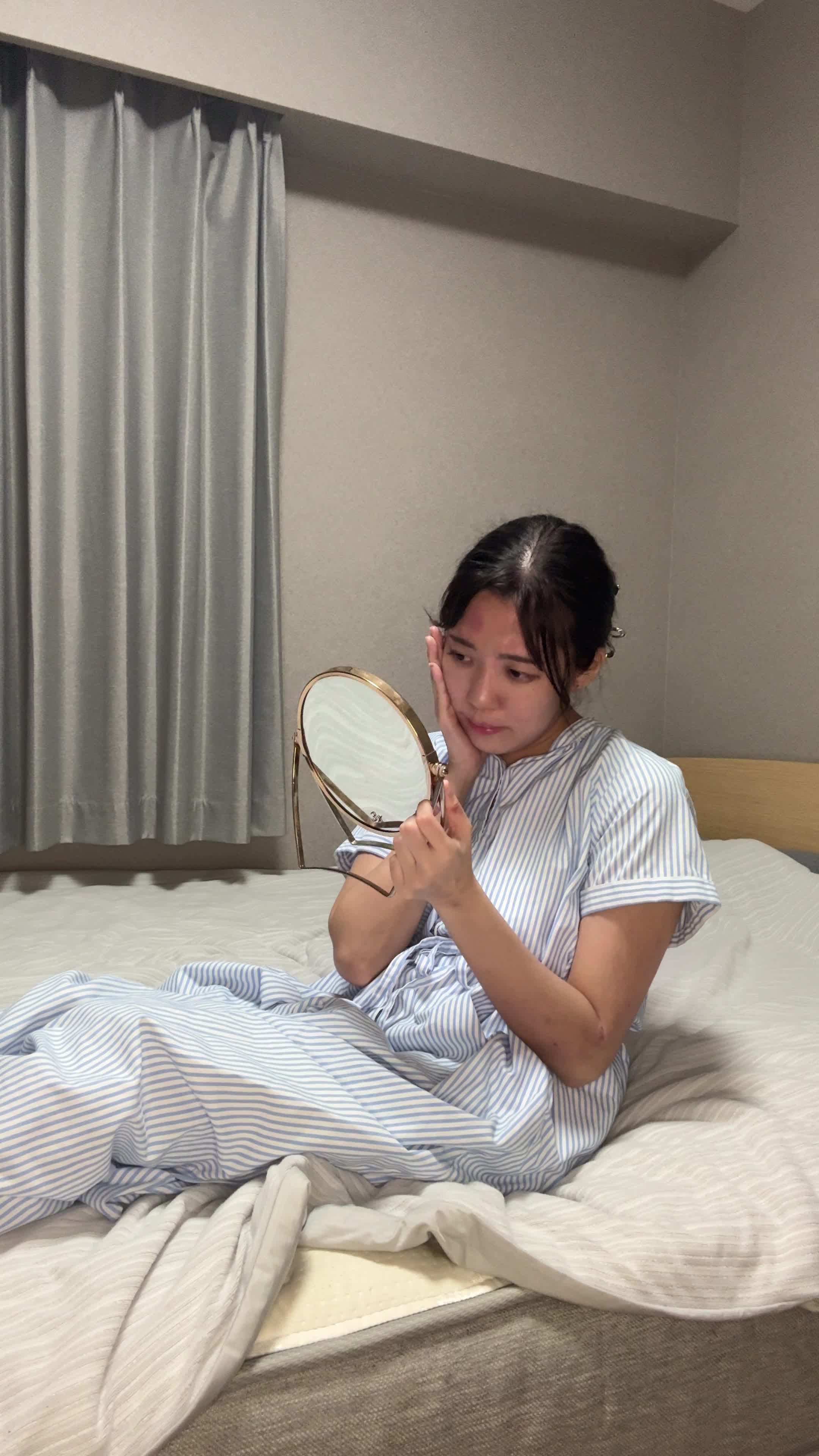 ベッドで脚を伸ばし、「便秘が原因で肌荒れしている気がする…」と鏡を見つめる動作