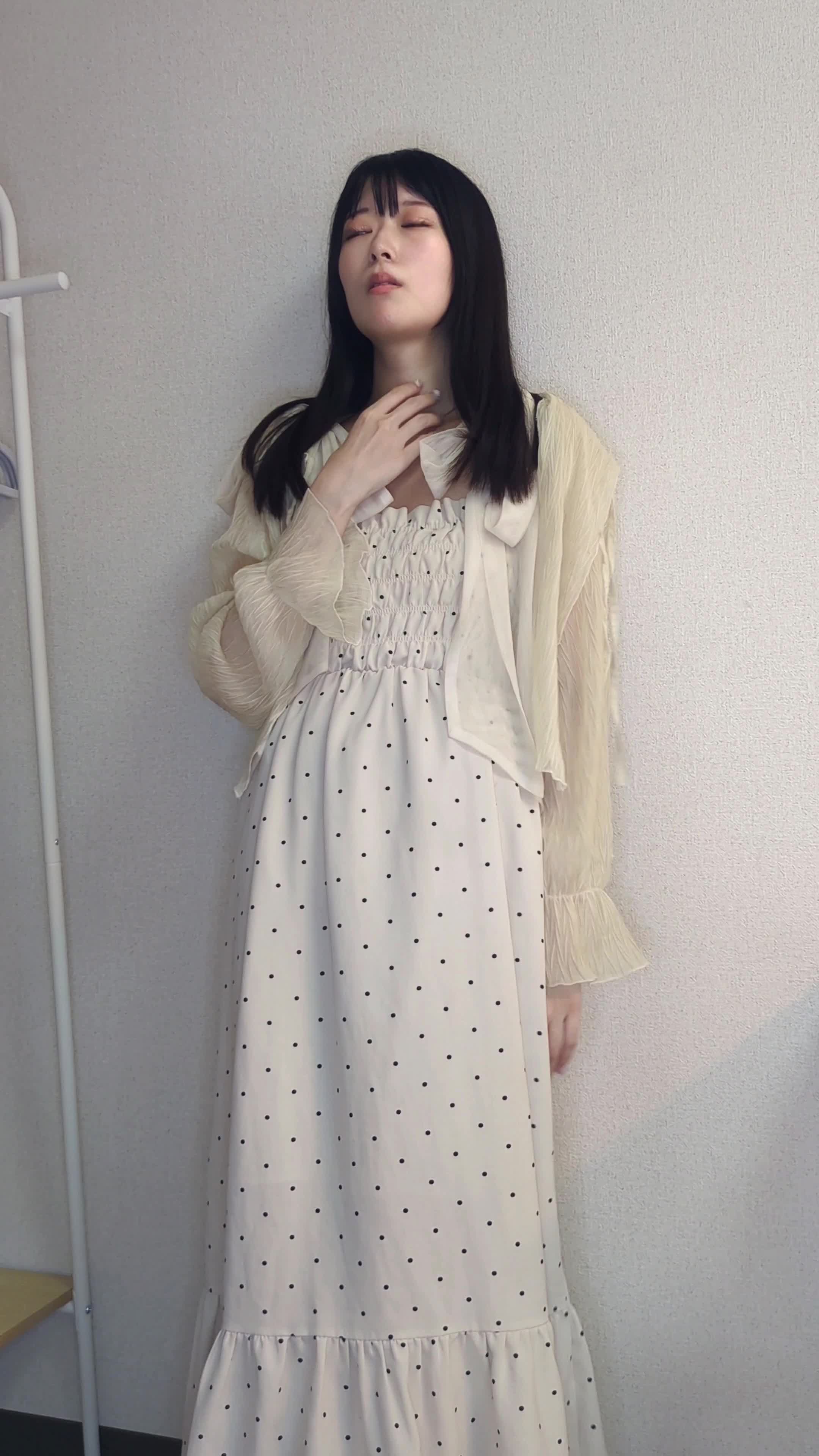 壁にもたれながら、手で軽く首元を押さえて肌を気にしている表情を見せる